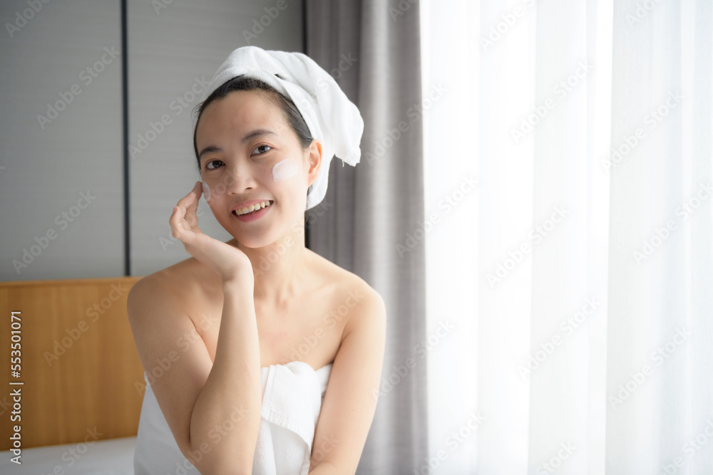 Happy young Asian woman applying face lotions while wearing a towel and touching her face. Daily makeup and skincare Happy young Asian woman applying face lotions while wearing a towel and touching her face. Daily makeup and skincare