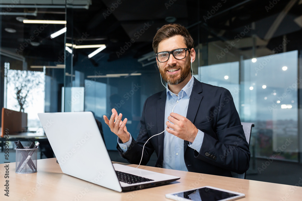A young businessman sits in the office at the table with headphones, conducts a business meeting, a conference through a video call from a laptop. He waves his hands, looks at the camera, smiles. A young businessman sits in the office at the table with headphones, conducts a business meeting, a conference through a video call from a laptop. He waves his hands, looks at the camera, smiles.