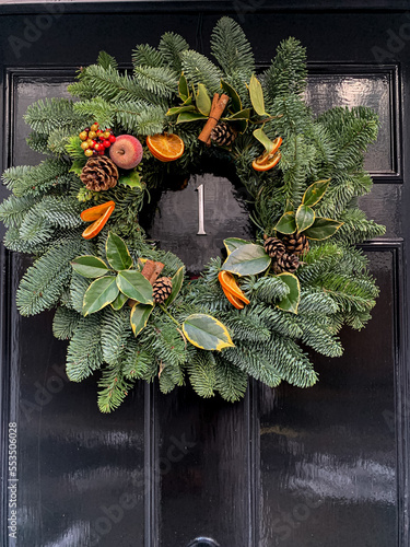 Selective focus of Christmas wreath. Festive christmasy themed winter natural wreath on a black wooden door, decorated with berries, pine cones, oranges, vanilla stick. Festive mood.