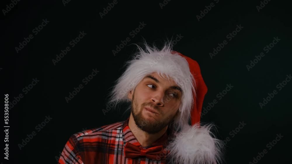 Lonely young Caucasian bearded man in red Santa hat wears plaid shirt and bow tie, portrait on dark studio background. Man sad, upset and depressed at Christmas holidays. Lonely young Caucasian bearded man in red Santa hat wears plaid shirt and bow tie, portrait on dark studio background. Man sad, upset and depressed at Christmas holidays.