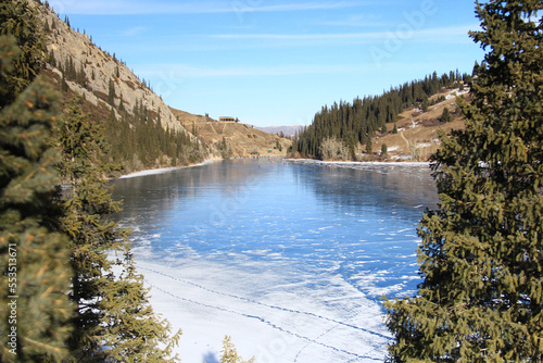 Alpine lake Kolsai covered with ice with a distant view of the mountain gorge, there is snow and traces on the ice in some places, spruce trees in the foreground, winter, sunny weather