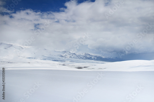 White snow-covered Assy plateau with mountain snow-covered ridges in the background, sunlight in the middle of the plateau and on the ridges, beautiful thick clouds, winter