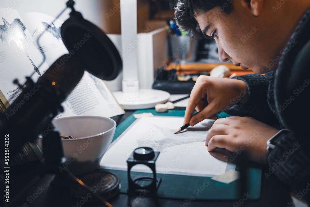 Close-up of a young man drawing at his study table. Young man holding a pencil and drawing something, warm orange light at home and a microphone for podcasts and social networking. Close-up of a young man drawing at his study table. Young man holding a pencil and drawing something, warm orange light at home and a microphone for podcasts and social networking.