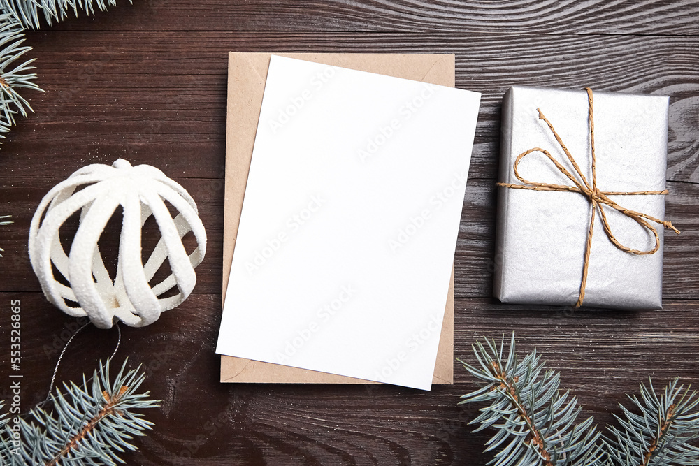 Christmas greeting card mockup with envelope, gift box, green fir tree branches and holiday decorations on brown wooden background, top view, flat lay. White New Year card with decor Christmas greeting card mockup with envelope, gift box, green fir tree branches and holiday decorations on brown wooden background, top view, flat lay. White New Year card with decor