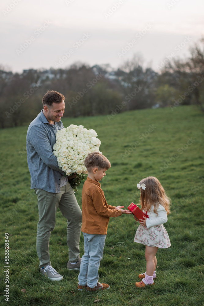 Fototapeta premium Happy little child boy gives smiling girl a gift box, making a surprise. Happy little girl is holding gift in red box and smiling, daddy holding flower
