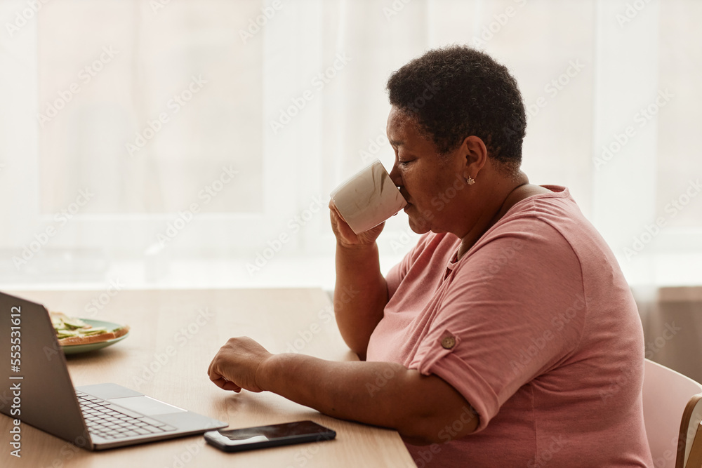Side view portrait of black senior woman enjoying cup of coffee at breakfast and using laptop Side view portrait of black senior woman enjoying cup of coffee at breakfast and using laptop