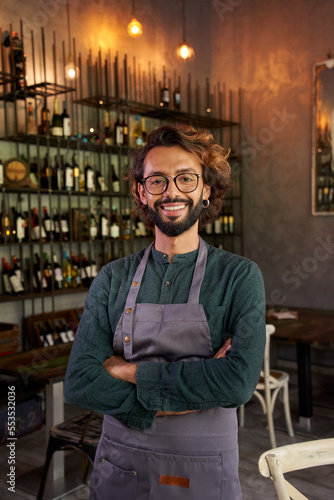 Looking at camera portrait of a young owner of a small local business. Cheerful male bartender smiling with positive attitude. Cheerful bar worker smiles while standing indoors.