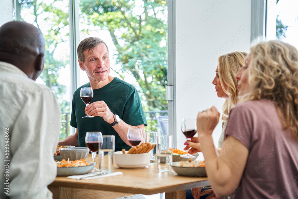 Group of fun mature diverse friends gathered at the table and raising their glasses in celebration Group of fun mature diverse friends gathered at the table and raising their glasses in celebration
