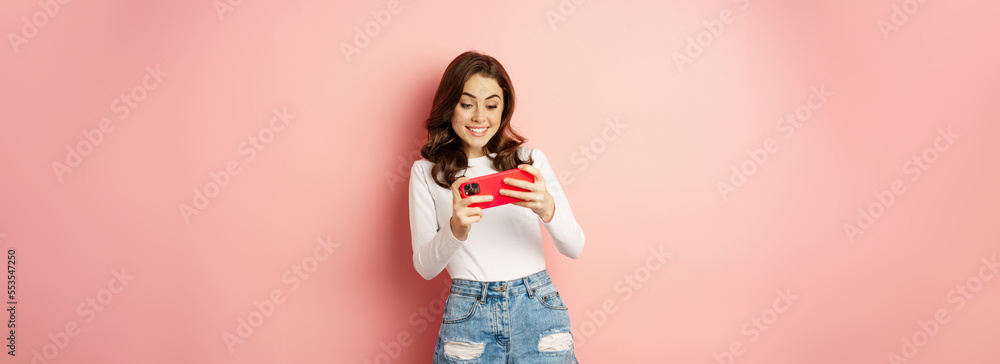 Free Photo  Happy beautiful girl playing mobile video game, holding  smartphone horizontally, watching on cellphone with excited face, pink  background.