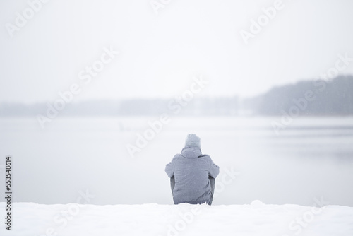 Obraz na płótnie Young adult man sitting alone on snow at lake shore and looking far away