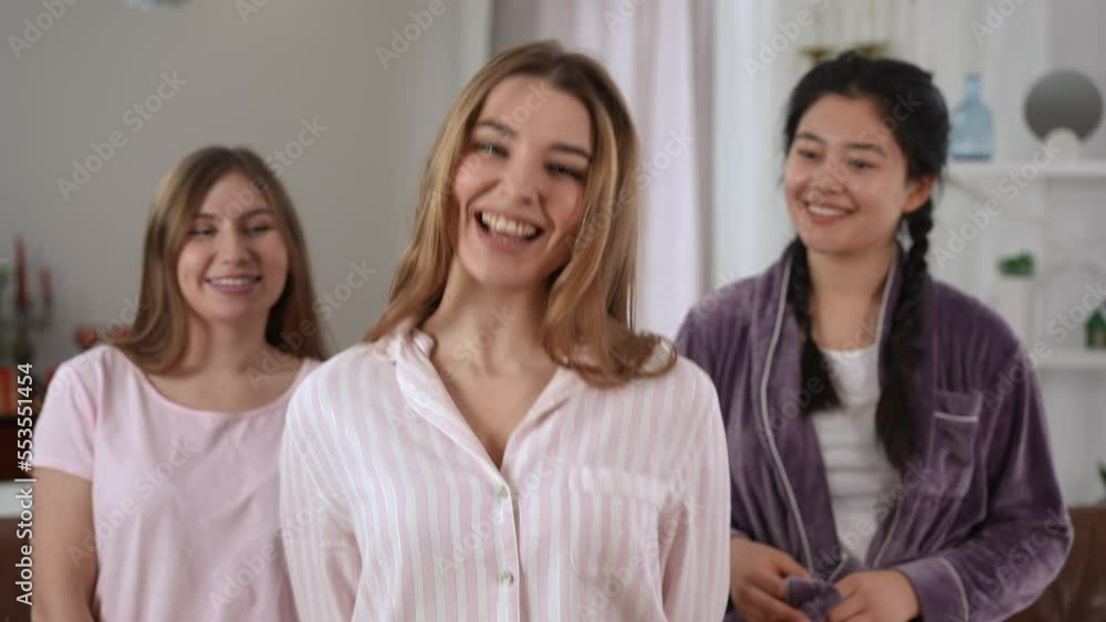 Portrait of happy gorgeous young woman looking at camera smiling with blurred friends standing at background. Relaxed confident Caucasian and Asian groupmates on pajama party posing Portrait of happy gorgeous young woman looking at camera smiling with blurred friends standing at background. Relaxed confident Caucasian and Asian groupmates on pajama party posing