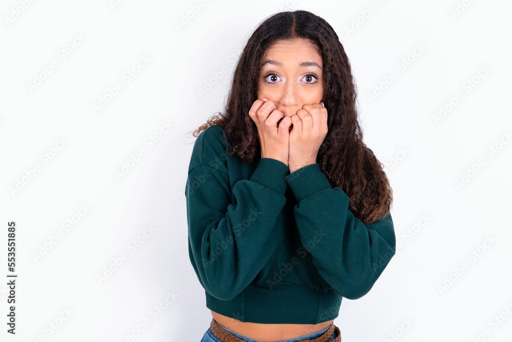 Anxiety - teen girl with curly hair wearing green sweater over white background covering his mouth with hands scared from something or someone bitting nails. Anxiety - teen girl with curly hair wearing green sweater over white background covering his mouth with hands scared from something or someone bitting nails.