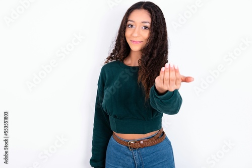 Bild auf Leinwand teen girl with curly hair wearing green sweater over white background, inviting you to come, confident and smiling making a gesture with hand, being positive and friendly