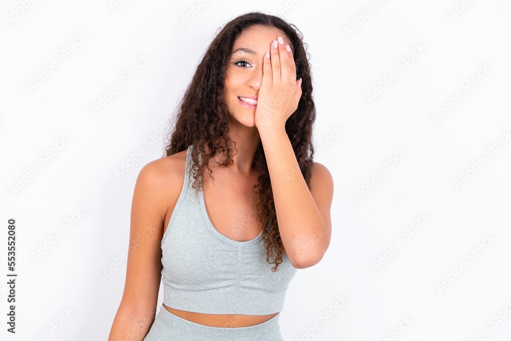 teen girl with curly hair wearing white sport set over gray background covering one eye with her hand, confident smile on face and surprise emotion. teen girl with curly hair wearing white sport set over gray background covering one eye with her hand, confident smile on face and surprise emotion.
