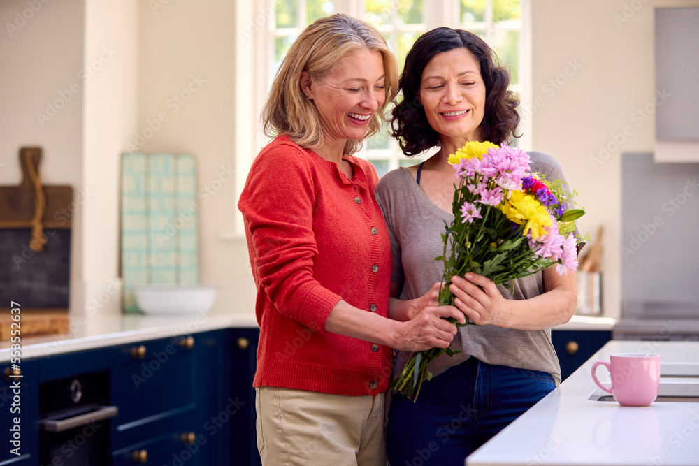 Loving Same Sex Mature Female Couple In Kitchen With Woman Giving Partner Flowers