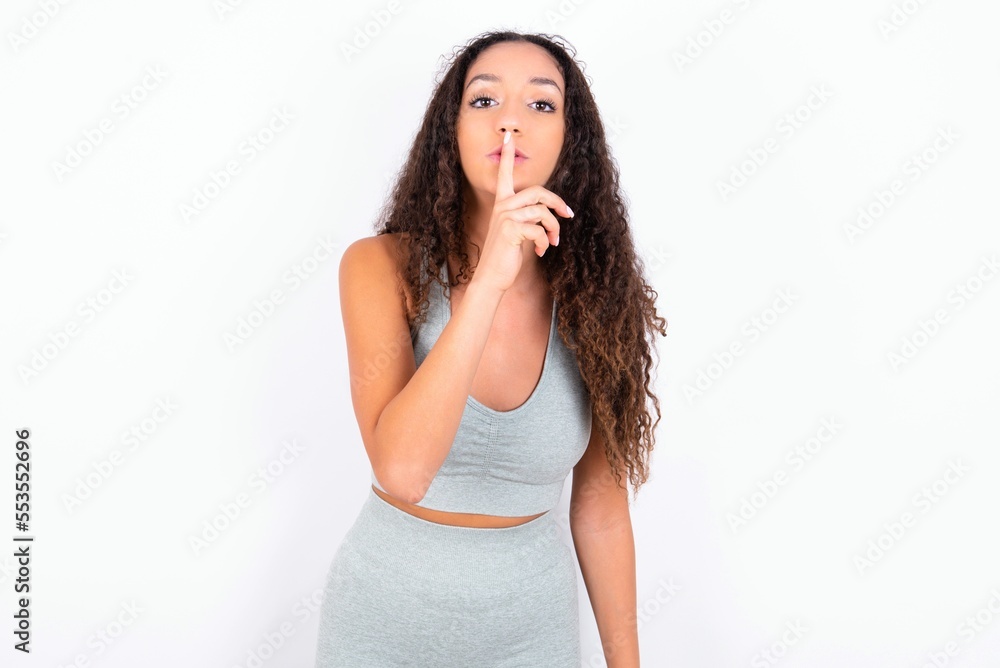 Beautiful teen girl with curly hair wearing grey sport set over white background makes silence gesture, keeps finger over lips. Silence and secret concept. Beautiful teen girl with curly hair wearing grey sport set over white background makes silence gesture, keeps finger over lips. Silence and secret concept.