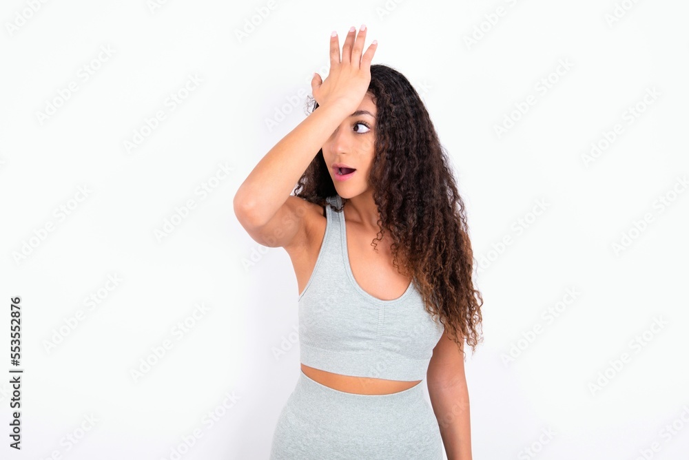Beautiful teen girl with curly hair wearing grey sport set over white background surprised with hand on head for mistake, remember error. Forgot, bad memory concept. Beautiful teen girl with curly hair wearing grey sport set over white background surprised with hand on head for mistake, remember error. Forgot, bad memory concept.