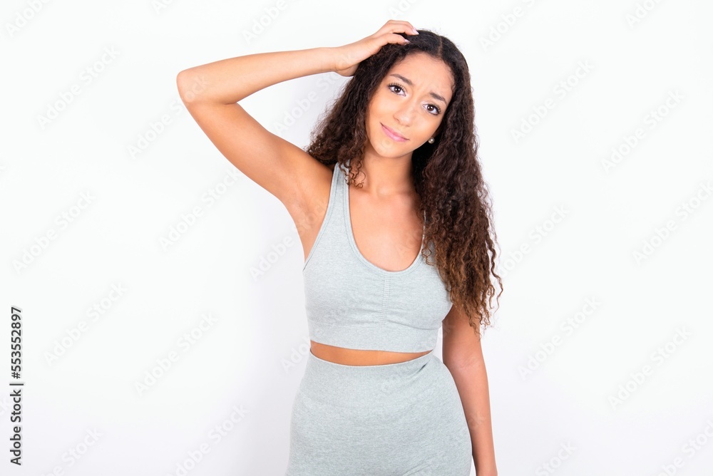 Beautiful teen girl with curly hair wearing grey sport set over white background being confused and wonders about something. Holding hand on head, uncertain with doubt. Pensive concept. Beautiful teen girl with curly hair wearing grey sport set over white background being confused and wonders about something. Holding hand on head, uncertain with doubt. Pensive concept.