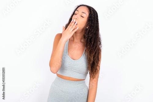 Beautiful teen girl with curly hair wearing grey sport set over white background being tired and yawning after spending all day at work. © Roquillo