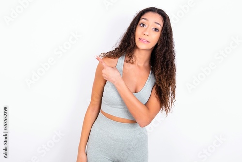 Beautiful teen girl with curly hair wearing grey sport set over white background Pointing aside worried and nervous with forefinger, concern and surprise concept.