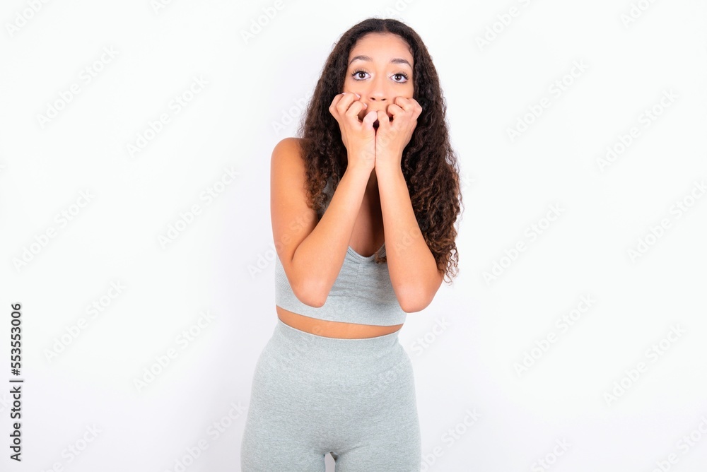 Fearful Beautiful teen girl with curly hair wearing grey sport set over white background keeps hands near mouth, feels frightened and scared, has a phobia, Shock and frighted concept. Fearful Beautiful teen girl with curly hair wearing grey sport set over white background keeps hands near mouth, feels frightened and scared, has a phobia, Shock and frighted concept.