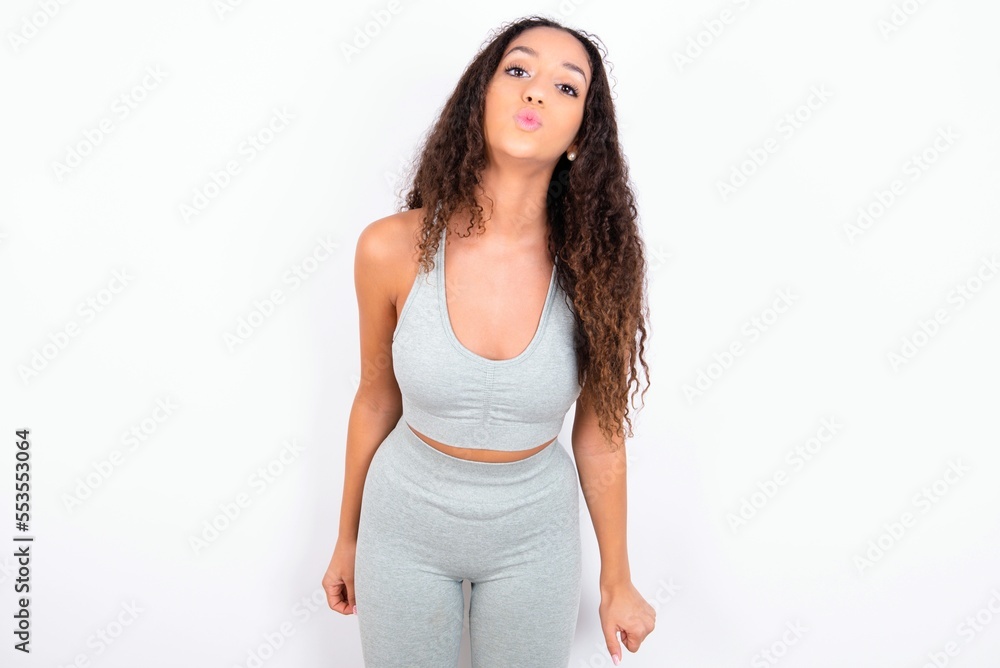 Shot of pleasant looking Beautiful teen girl with curly hair wearing grey sport set over white background, pouts lips, looks at camera, Human facial expressions Shot of pleasant looking Beautiful teen girl with curly hair wearing grey sport set over white background, pouts lips, looks at camera, Human facial expressions