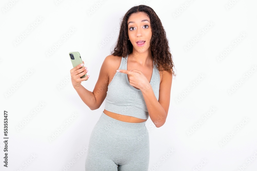 Beautiful teen girl with curly hair wearing green sport set over white background holding in hands showing new cell, Beautiful teen girl with curly hair wearing green sport set over white background holding in hands showing new cell,