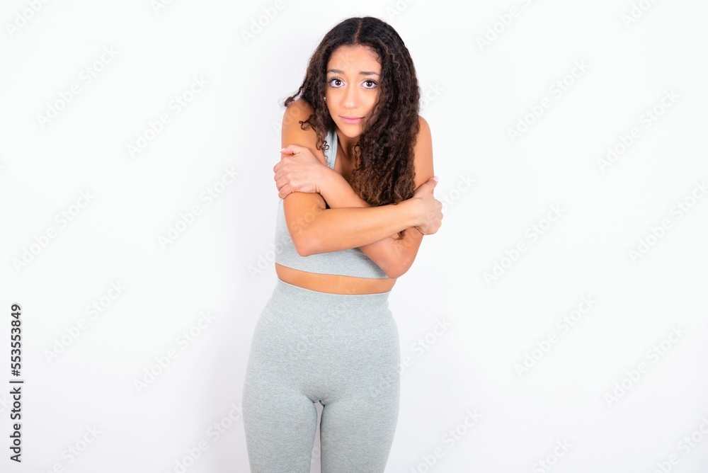 Desperate Beautiful teen girl with curly hair wearing green sport set over white background trembles and feels cold, hugs oneself to warm up or feels scared notices something terrifying.