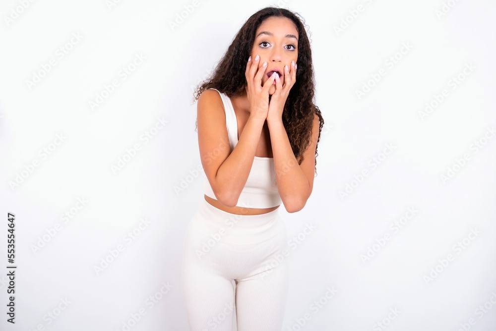 Speechless teen girl with curly hair wearing white sport set over white background keeps hands near opened mouth reacts to shocking news stares wondered at camera Speechless teen girl with curly hair wearing white sport set over white background keeps hands near opened mouth reacts to shocking news stares wondered at camera