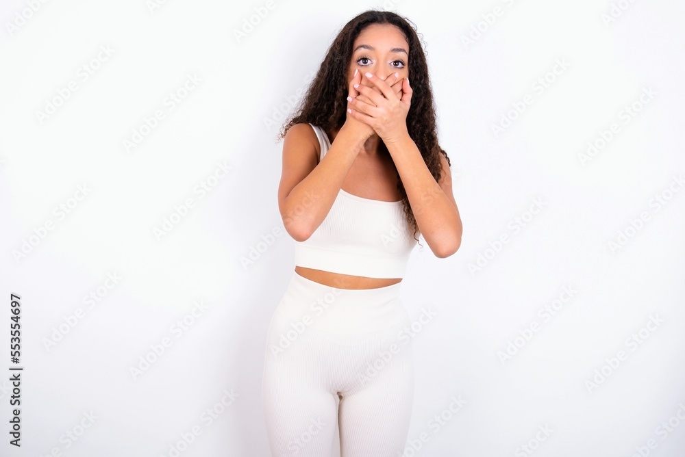 Stunned teen girl with curly hair wearing white sport set over white background covers both hands on mouth, afraids of something astonishing Stunned teen girl with curly hair wearing white sport set over white background covers both hands on mouth, afraids of something astonishing