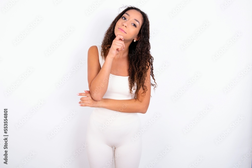 teen girl with curly hair wearing white sport set over white background looking confident at the camera smiling with crossed arms and hand raised on chin. Thinking positive. teen girl with curly hair wearing white sport set over white background looking confident at the camera smiling with crossed arms and hand raised on chin. Thinking positive.
