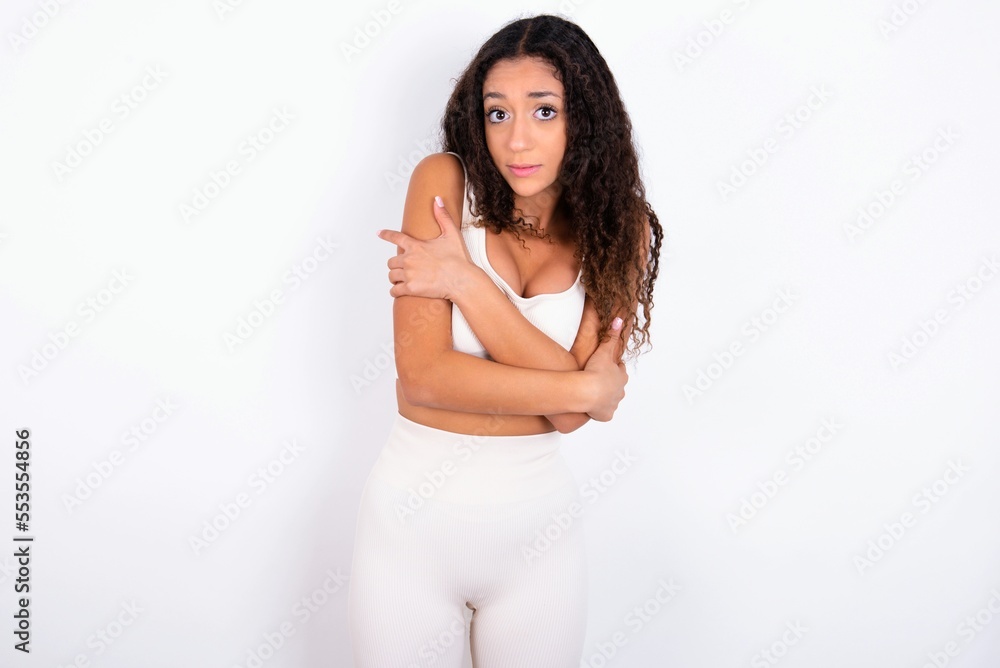 teen girl with curly hair wearing white sport set over white background shaking and freezing for winter cold with sad and shock expression on face. teen girl with curly hair wearing white sport set over white background shaking and freezing for winter cold with sad and shock expression on face.