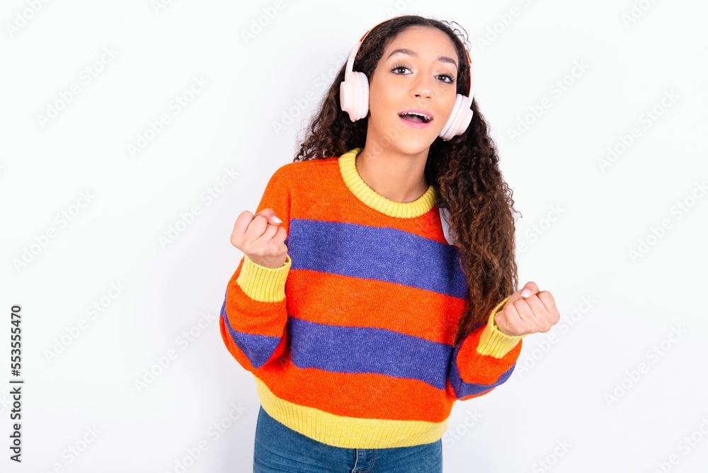 Emotional beautiful teen girl wearing sweater exclaims loudly feels like winner raises clenched fists keeps mouth opened wears stereo headphones on ears makes yes gesture, listens favourite music Emotional beautiful teen girl wearing sweater exclaims loudly feels like winner raises clenched fists keeps mouth opened wears stereo headphones on ears makes yes gesture, listens favourite music