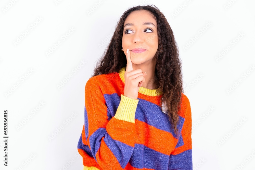 Lovely dreamy beautiful teen girl wearing striped knitted sweater over white background keeps finger near lips looks aside copy space. Lovely dreamy beautiful teen girl wearing striped knitted sweater over white background keeps finger near lips looks aside copy space.