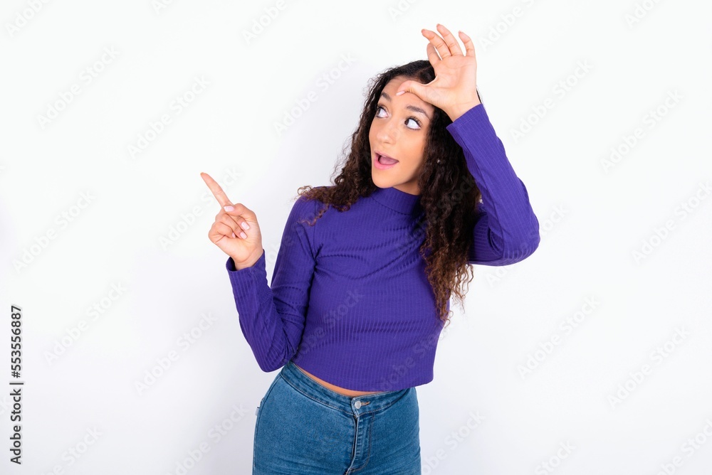 beautiful teen girl wearing knitted purple sweater over white background showing loser sign and pointing at empty space beautiful teen girl wearing knitted purple sweater over white background showing loser sign and pointing at empty space