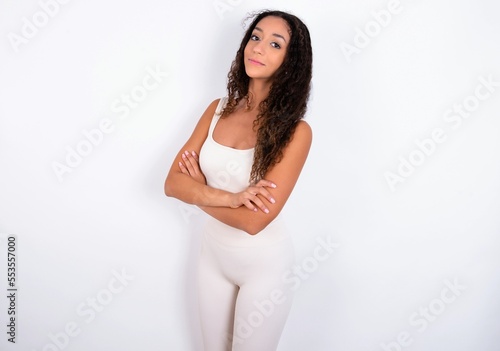 Portrait of teen girl with curly hair wearing white sport set over white background standing with folded arms and smiling © Roquillo