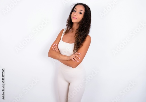 Portrait of teen girl with curly hair wearing white sport set over white background standing with folded arms and smiling