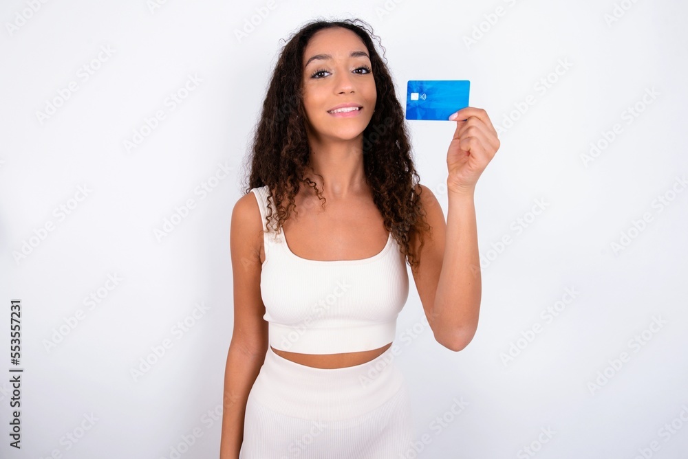 Photo of happy cheerful smiling positive teen girl with curly hair wearing white sport set over white background recommend credit card Photo of happy cheerful smiling positive teen girl with curly hair wearing white sport set over white background recommend credit card