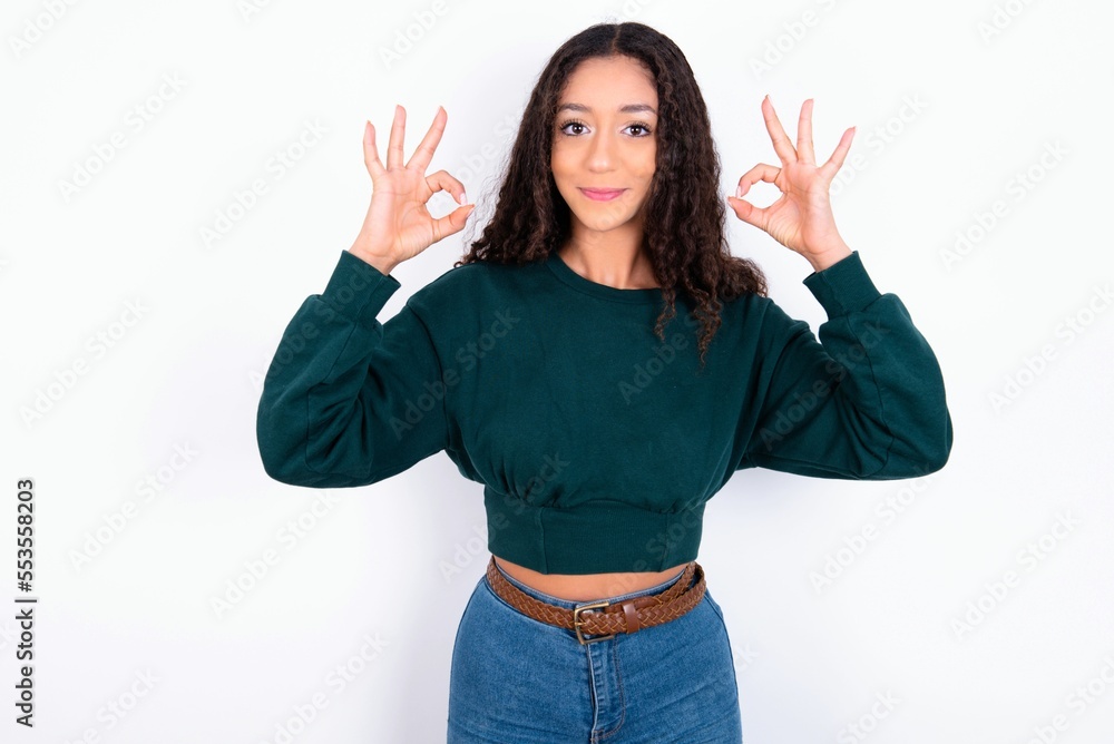 Glad teen girl wearing knitted green sweater over white background shows ok sign with both hands as expresses approval, has cheerful expression, being optimistic. Glad teen girl wearing knitted green sweater over white background shows ok sign with both hands as expresses approval, has cheerful expression, being optimistic.