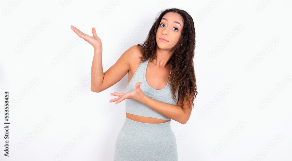 teen girl with curly hair wearing white sport set over gray background pointing aside with both hands showing something strange and saying: I don't know what is this. Advertisement concept. teen girl with curly hair wearing white sport set over gray background pointing aside with both hands showing something strange and saying: I don't know what is this. Advertisement concept.