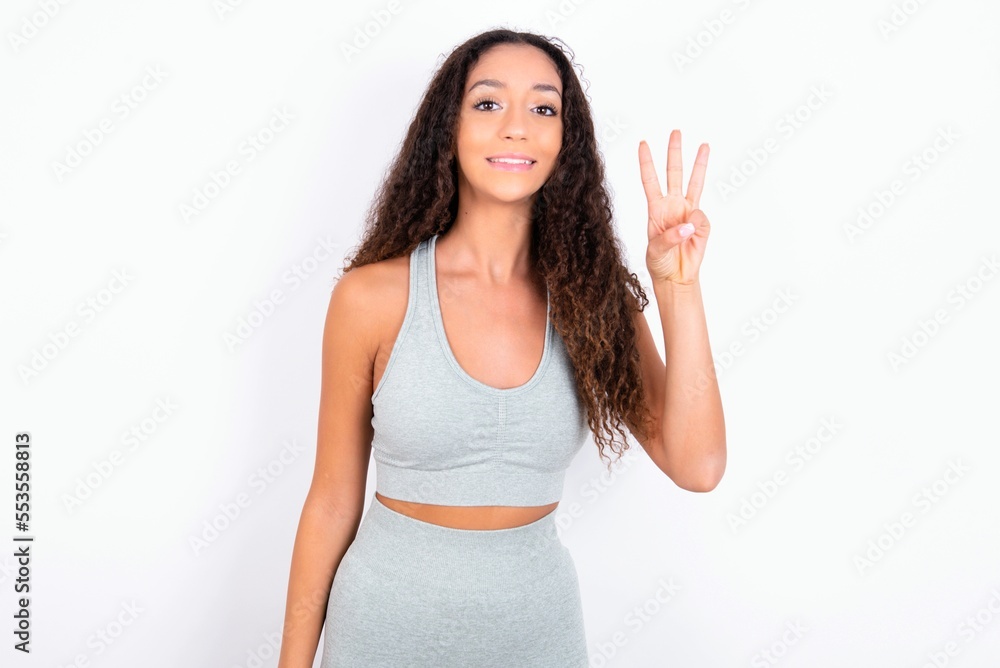 teen girl with curly hair wearing white sport set over gray background showing and pointing up with fingers number three while smiling confident and happy. teen girl with curly hair wearing white sport set over gray background showing and pointing up with fingers number three while smiling confident and happy.