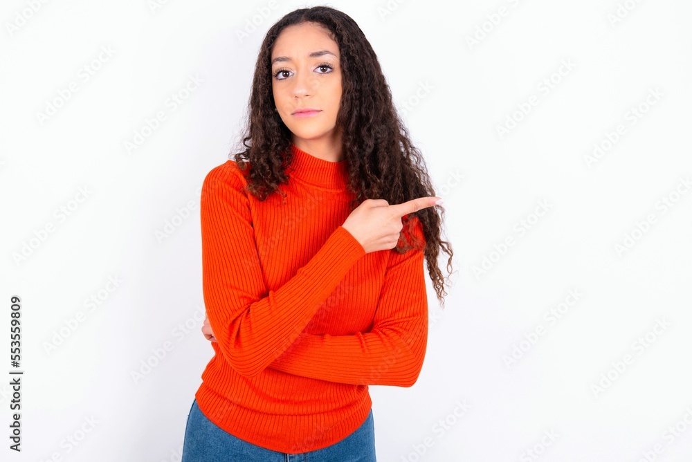 teen girl wearing knitted red sweater over white background smiling broadly at camera, pointing fingers away, showing something interesting and exciting. teen girl wearing knitted red sweater over white background smiling broadly at camera, pointing fingers away, showing something interesting and exciting.