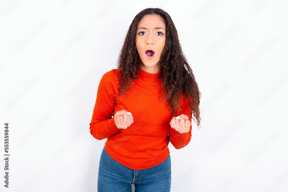 Joyful excited lucky teen girl wearing knitted red sweater over white background cheering, celebrating success, screaming yes with clenched fists Joyful excited lucky teen girl wearing knitted red sweater over white background cheering, celebrating success, screaming yes with clenched fists