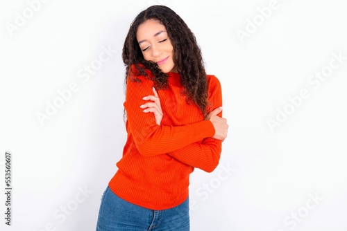 teen girl wearing knitted red sweater over white background . Hugging oneself happy and positive, smiling confident. Self love and self care. © Jihan