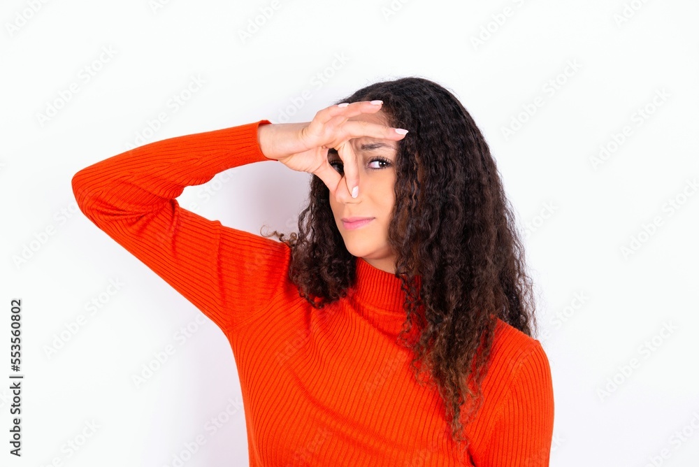 Displeased teen girl wearing knitted red sweater over white background plugs nose as smells something stink and unpleasant feels aversion hates disgusting scent. Stock Photo Adobe Stock