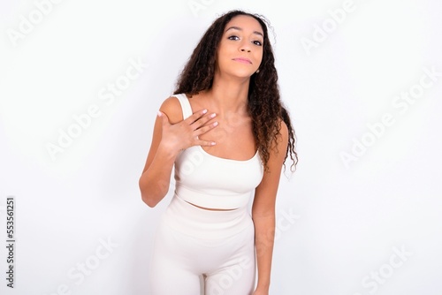 teen girl with curly hair wearing white sport set over white background smiles toothily cannot believe eyes expresses good emotions and surprisement