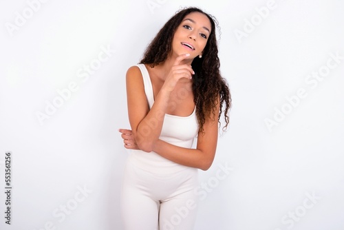 teen girl with curly hair wearing white sport set over white background laughs happily keeps hand on chin expresses positive emotions smiles broadly has carefree expression