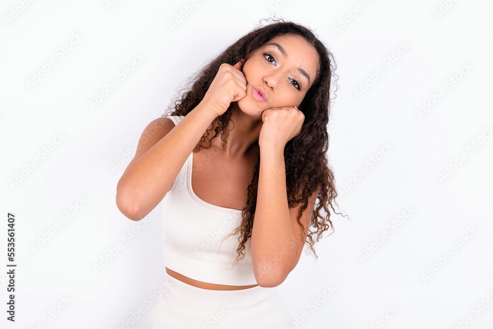 teen girl with curly hair wearing white sport set over white background with surprised expression keeps hands under chin keeps lips folded makes funny grimace