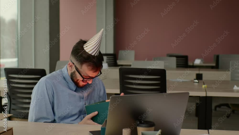 Happy positive bearded office worker in cone hat sitting at workplace with laptop and gift with festive ribbon. Happy positive bearded office worker in cone hat sitting at workplace with laptop and gift with festive ribbon.