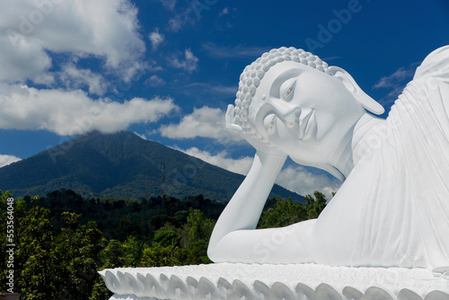 White sleeping buddha statue in a reclining pose with a mountain and forest in the background, Vihara Dharma Giri Temple; Tabanan, Bali, Indonesia photo